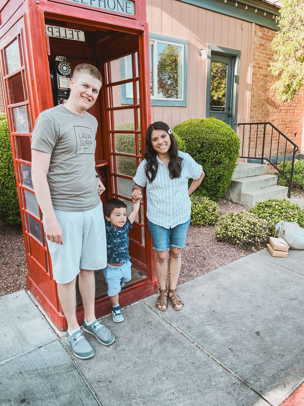 mom and dad holding hands with small dark-haired boy in front of a red telephone booth for family travel blog